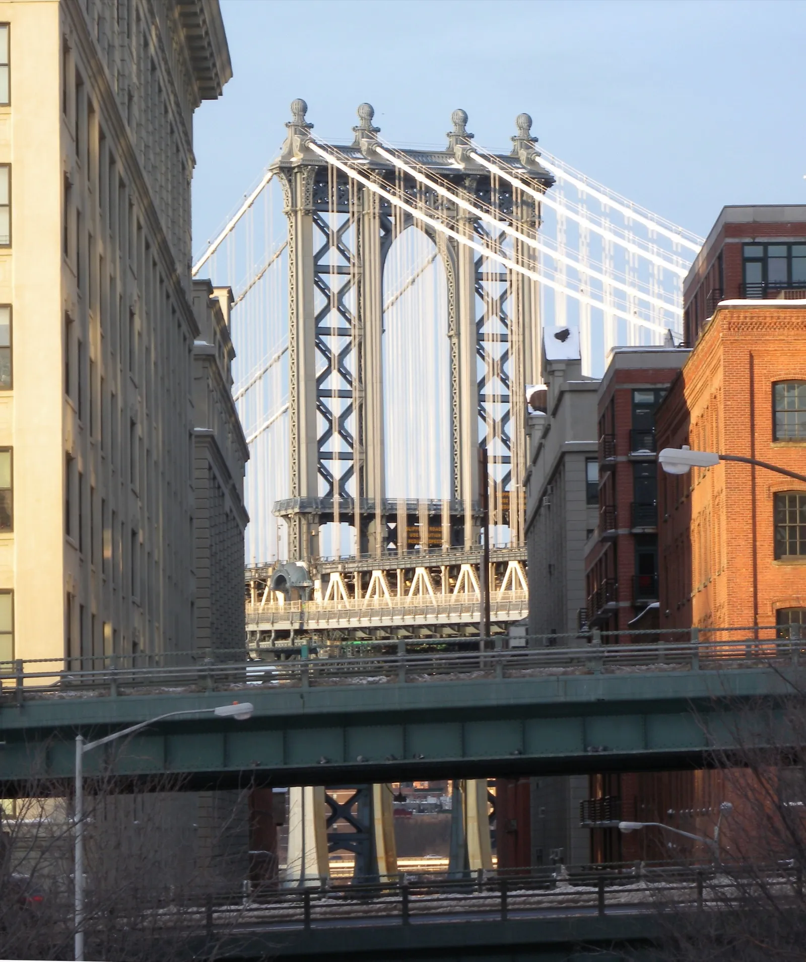 The steel tower of the Manhattan Bridge rises above Washington Street in DUMBO Brooklyn framed by brick warehouse buildings and an elevated roadway in the foreground