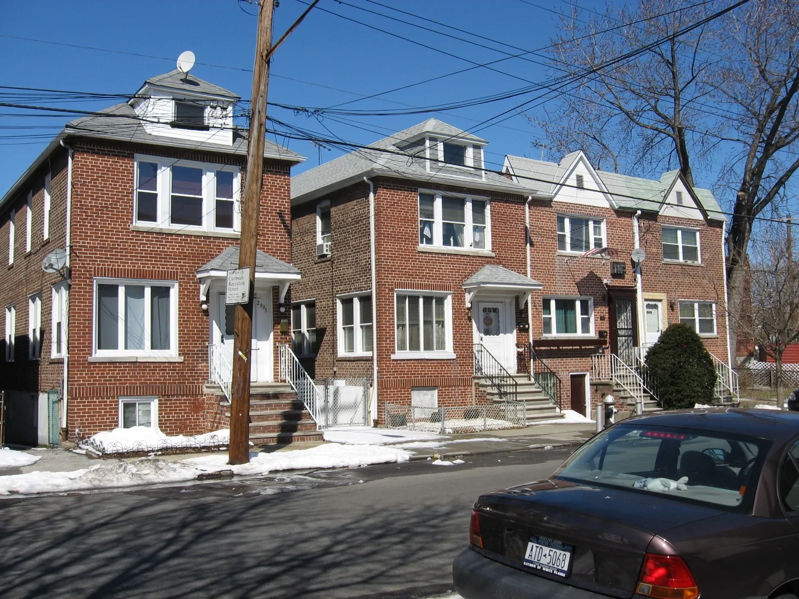 Attached brick two-family houses on a residential street in Pelham Bay, Bronx, with a car parked at the curb
