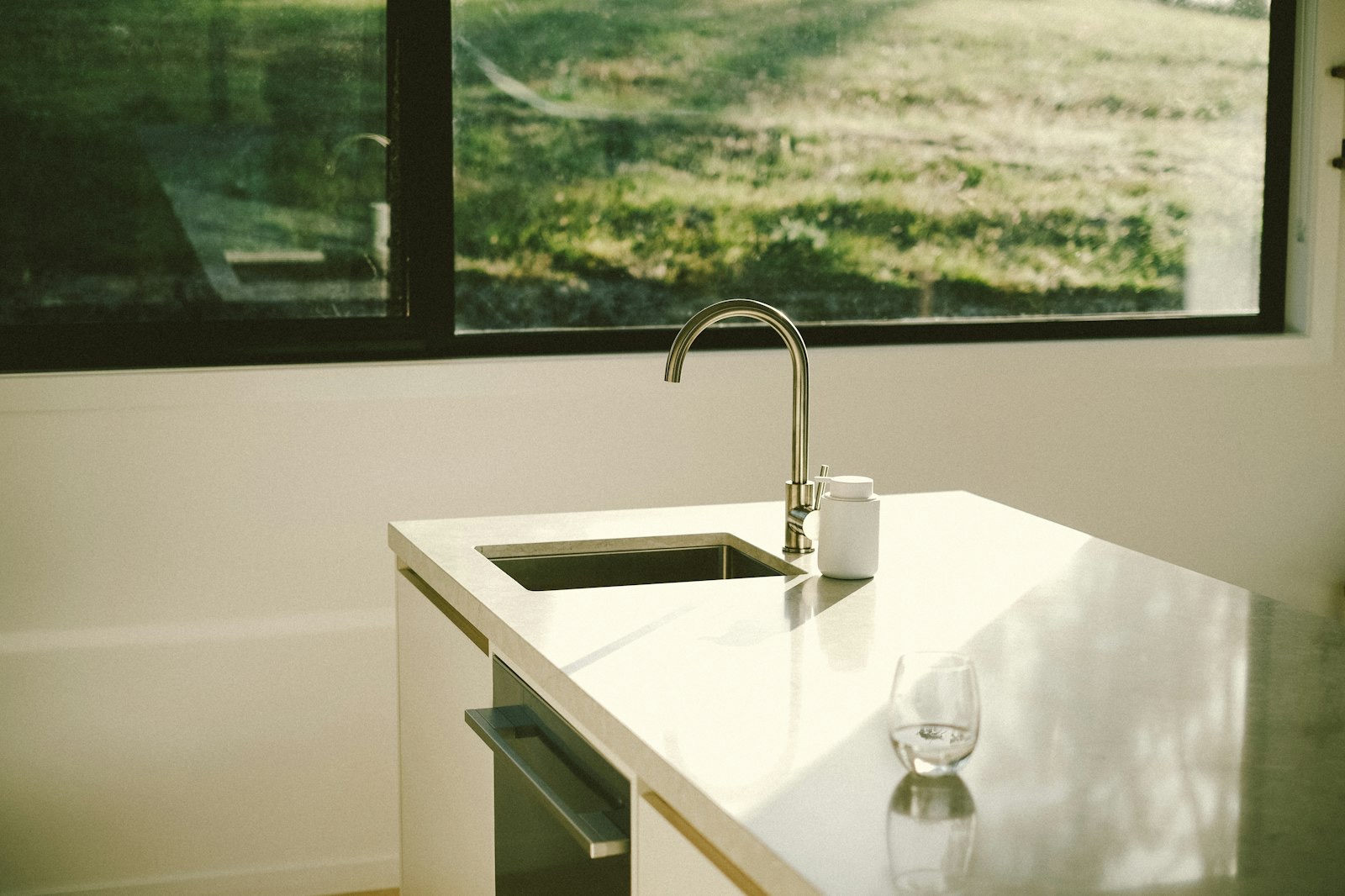 A cleared stone kitchen counter with an undermount sink and a single glass, lit by low sun through the window