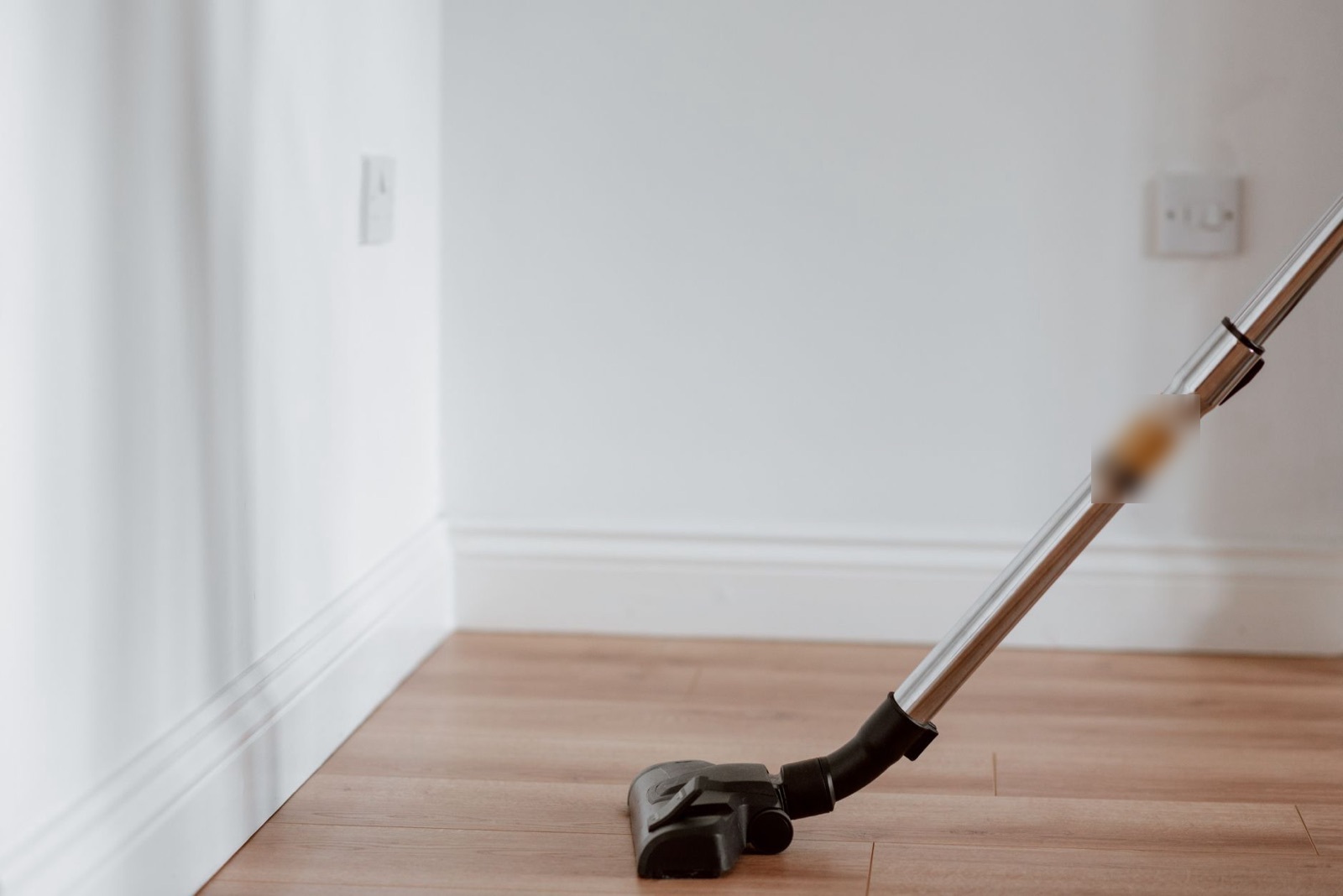 A vacuum cleaner head and chrome wand on light wood floorboards in an empty room with white walls