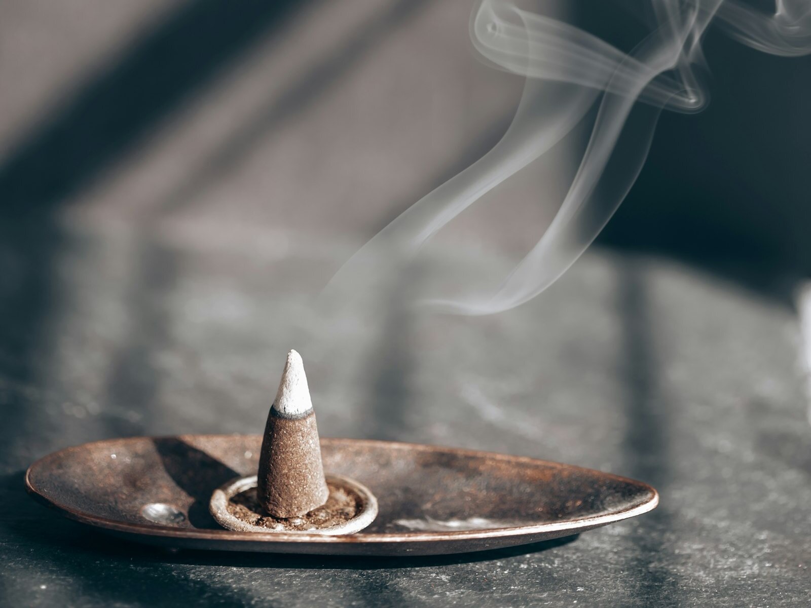 An incense cone burning on a metal dish in window light, with white ash at its tip and gray ash collected in the ring around its base