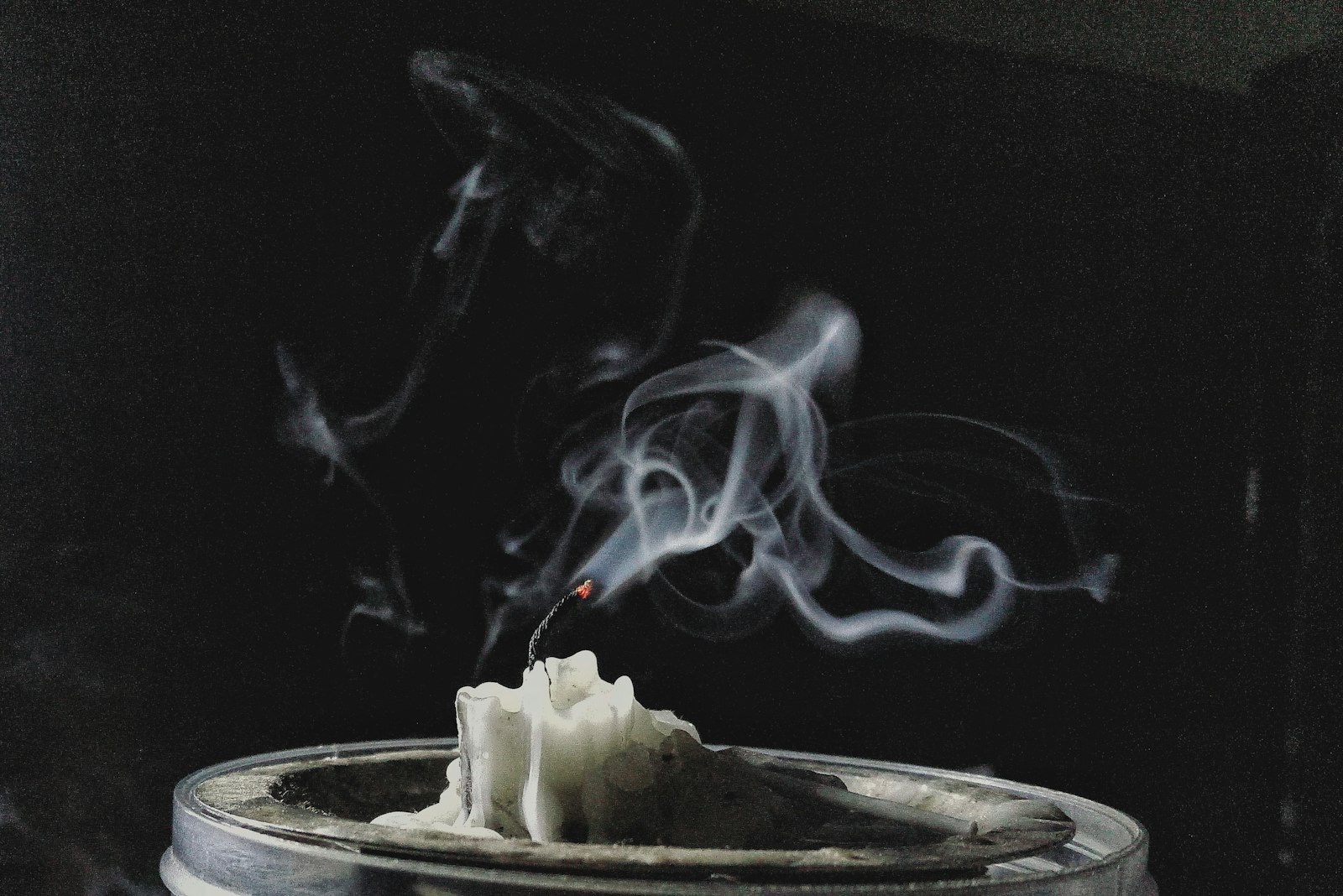 A burned-down candle just blown out, its long wick still glowing and trailing smoke, with dark soot residue on the metal lid around the wax