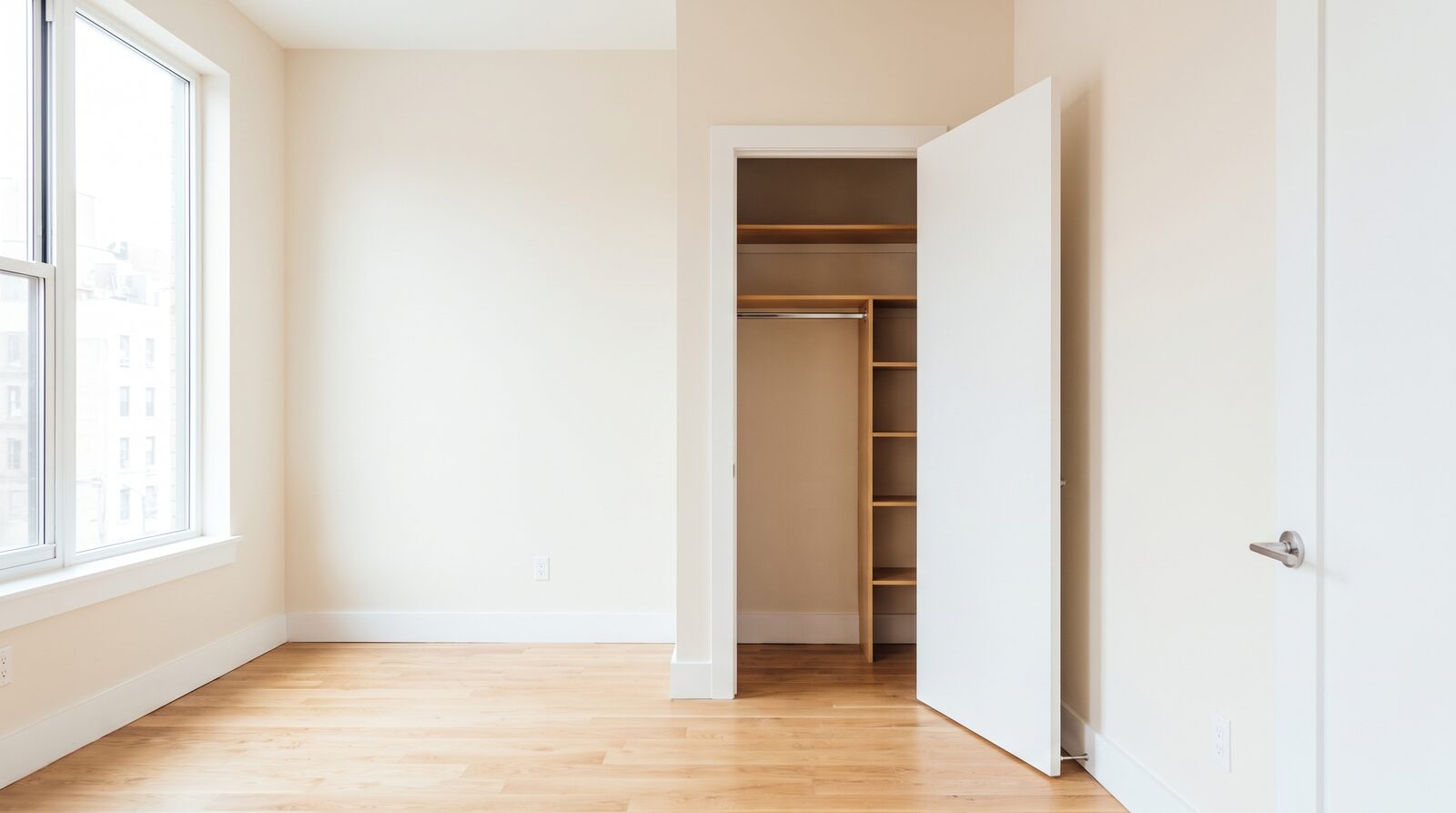 Empty NYC apartment bedroom after a move-out clean, with bare warm-toned walls, a swept and mopped oak floor, and the closet door open on emptied shelves and a bare hanging rod