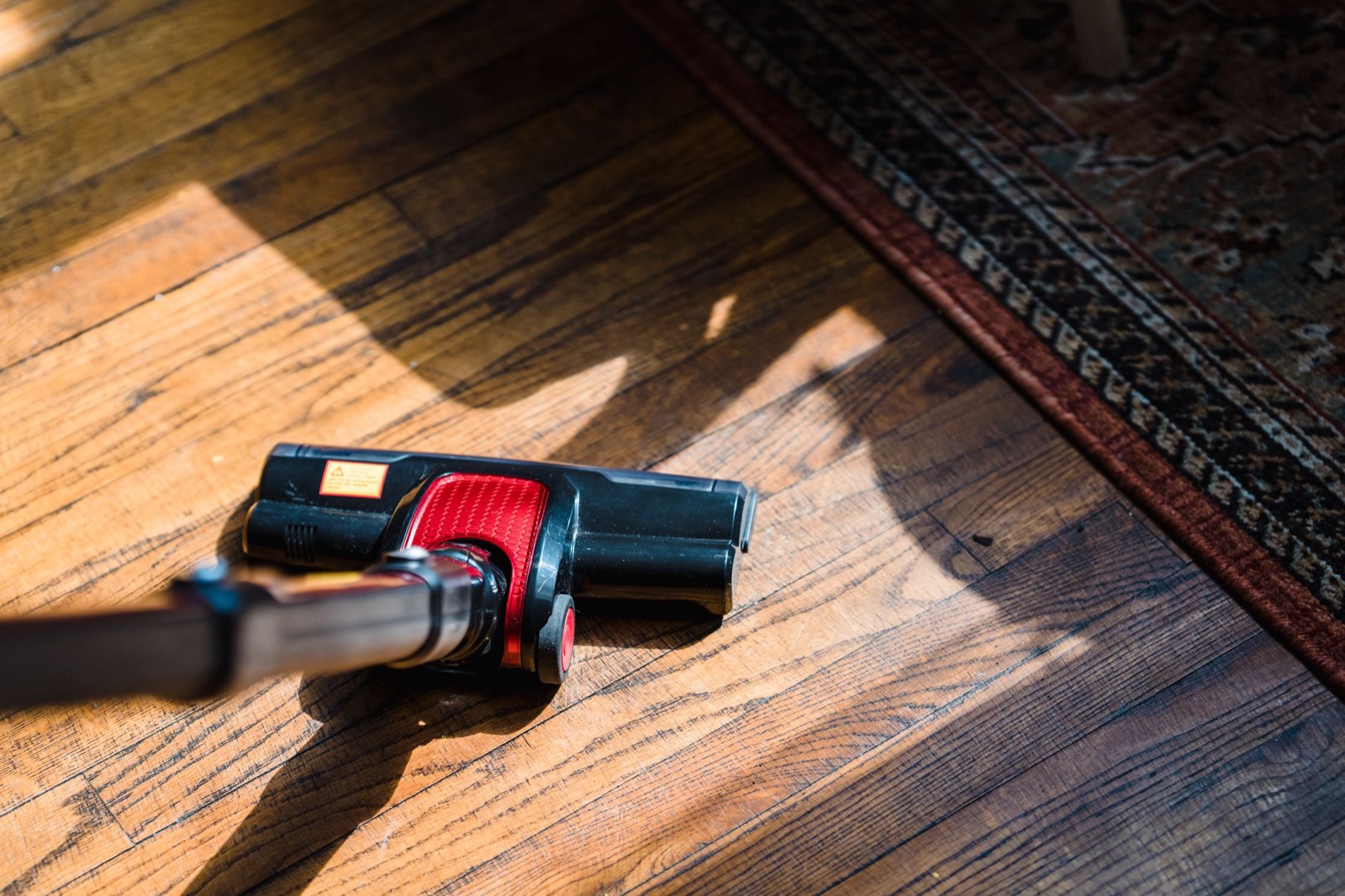 A black and red vacuum head resting on sunlit oak strip flooring beside the edge of a patterned rug