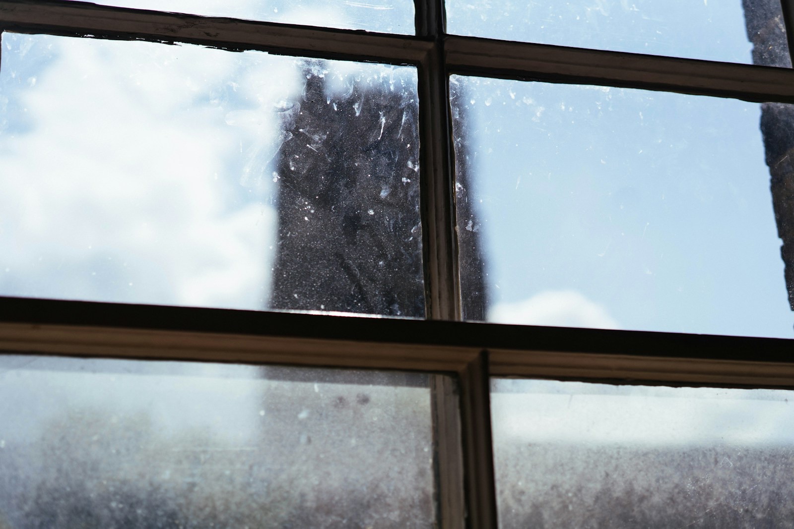 Bright sunlight catching fine curved marks and specks on the glass of an old multi-pane window, a dark stone tower and blue sky beyond