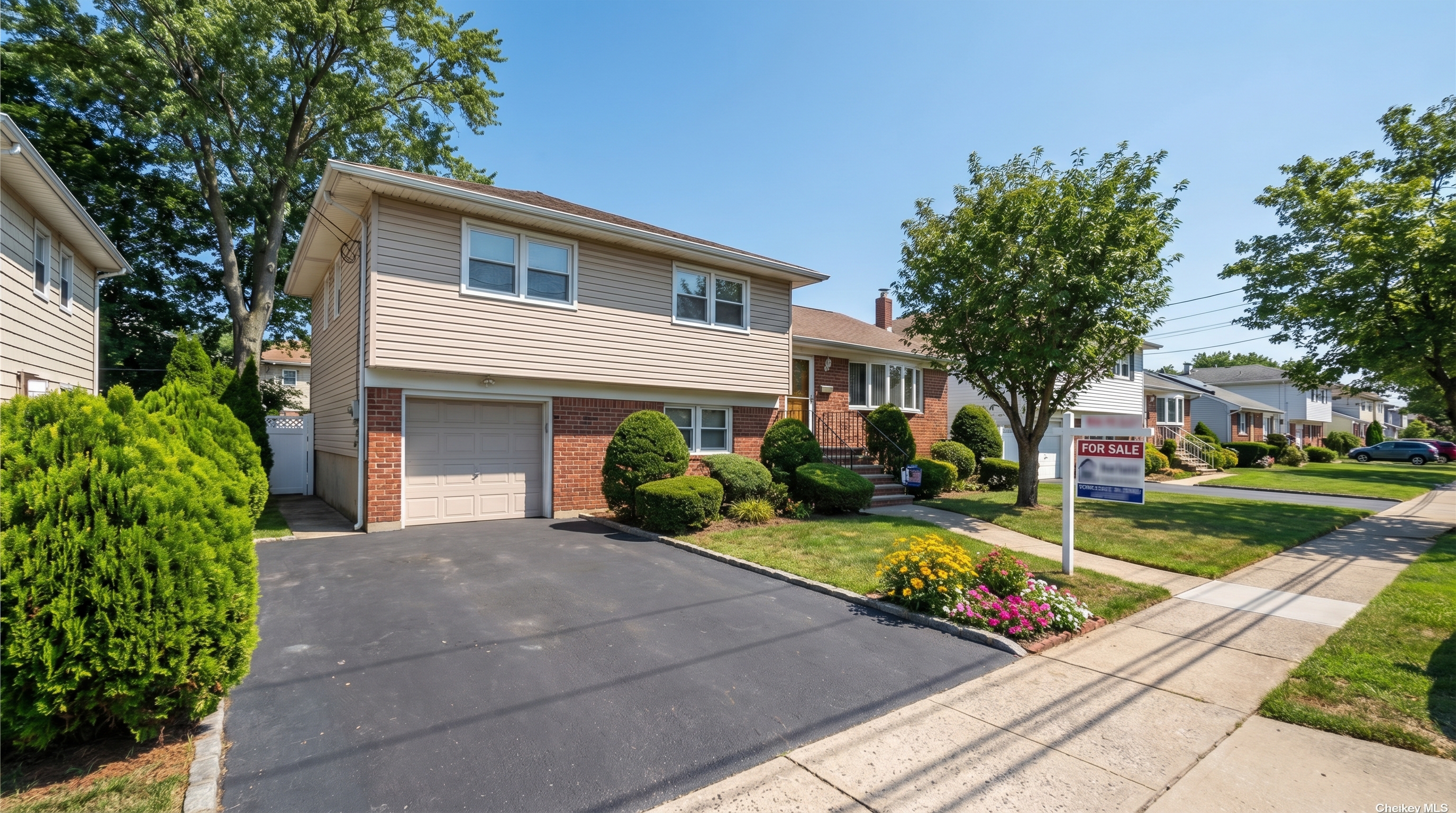 A postwar split-level home in southeastern Queens with a brick lower level, aluminum siding upper level, attached garage, and carefully manicured front garden with flowering plants