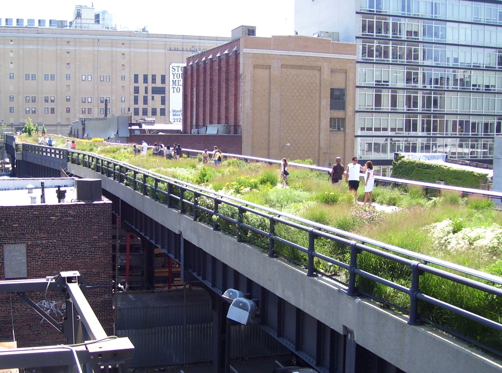 The High Line elevated park near 20th Street, showing visitors walking along the converted rail line surrounded by lush green plantings, with industrial buildings and apartment towers flanking both sides