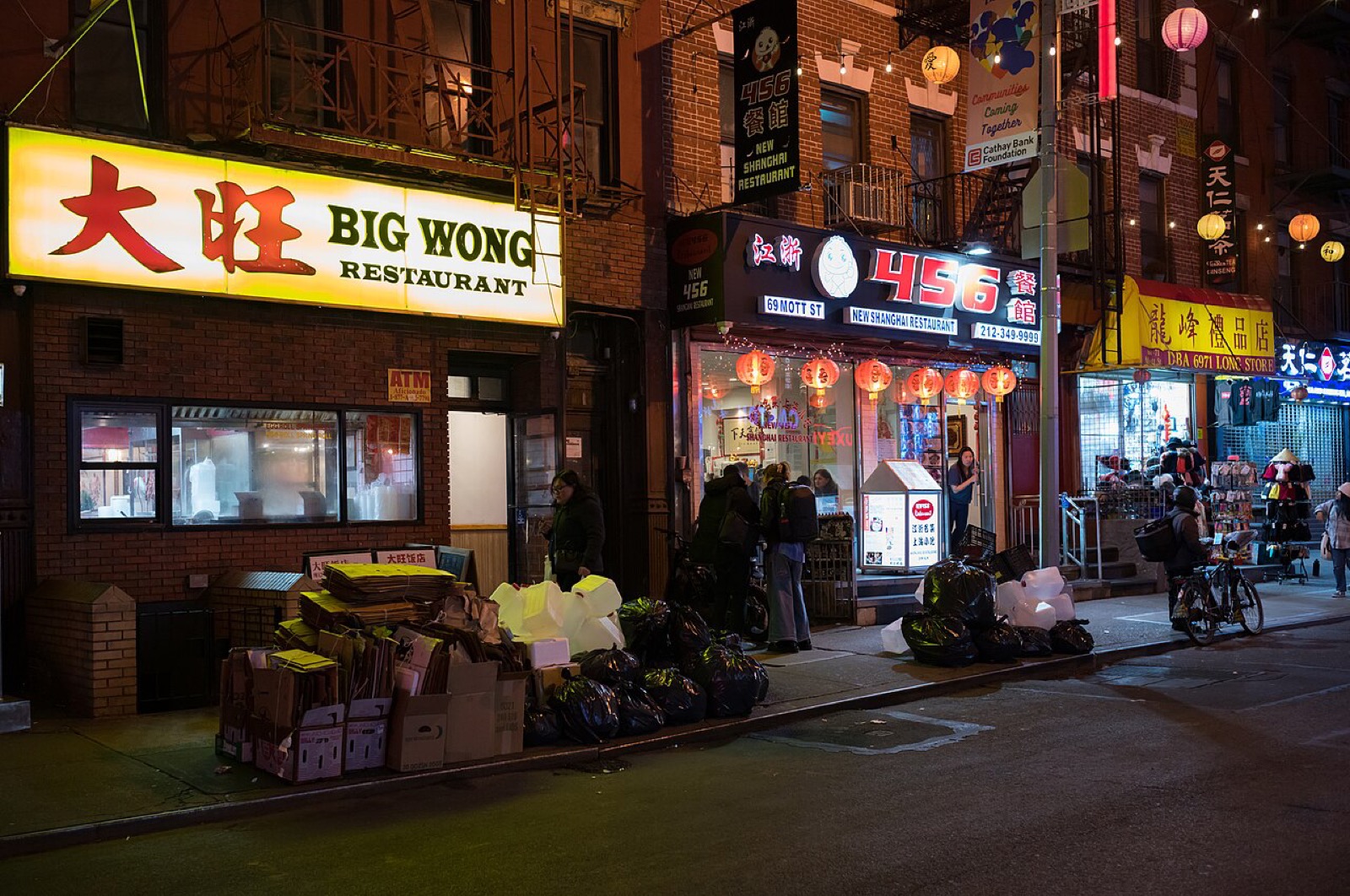 Mott Street in Chinatown lined with Chinese storefronts, restaurant signs in Chinese and English, and pedestrians walking along the narrow sidewalks