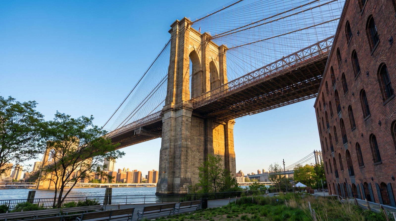 The Brooklyn Bridge's Gothic stone towers rising above Fulton Landing Park in DUMBO, with suspension cables radiating outward, the red brick Empire Stores warehouse building to the right, and park greenery in the foreground under a clear sky