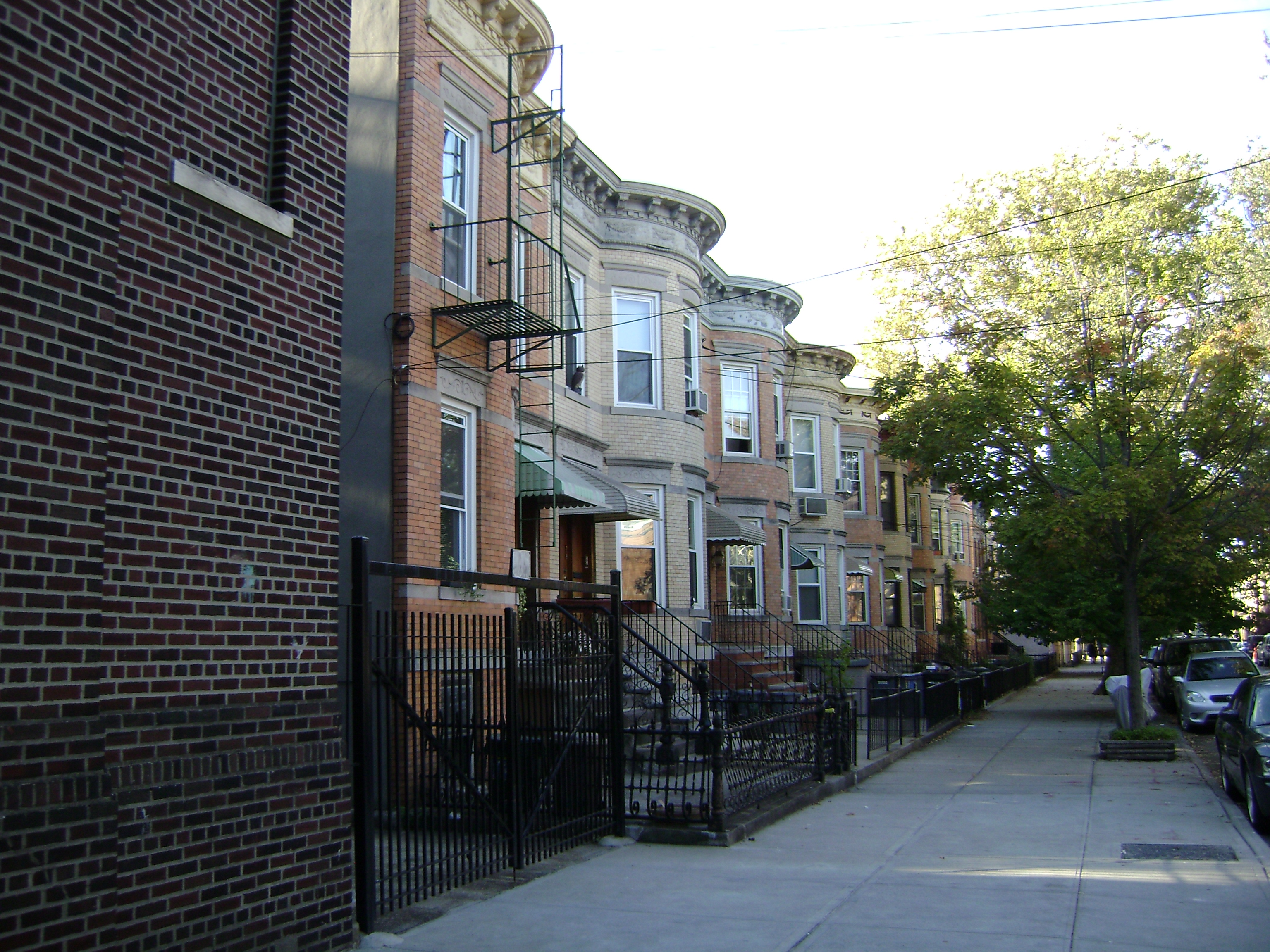 Brick two-family row houses on a Glendale Queens residential block showing attached facades, small front gardens, and maintained stoops characteristic of the 1910s to 1930s housing stock