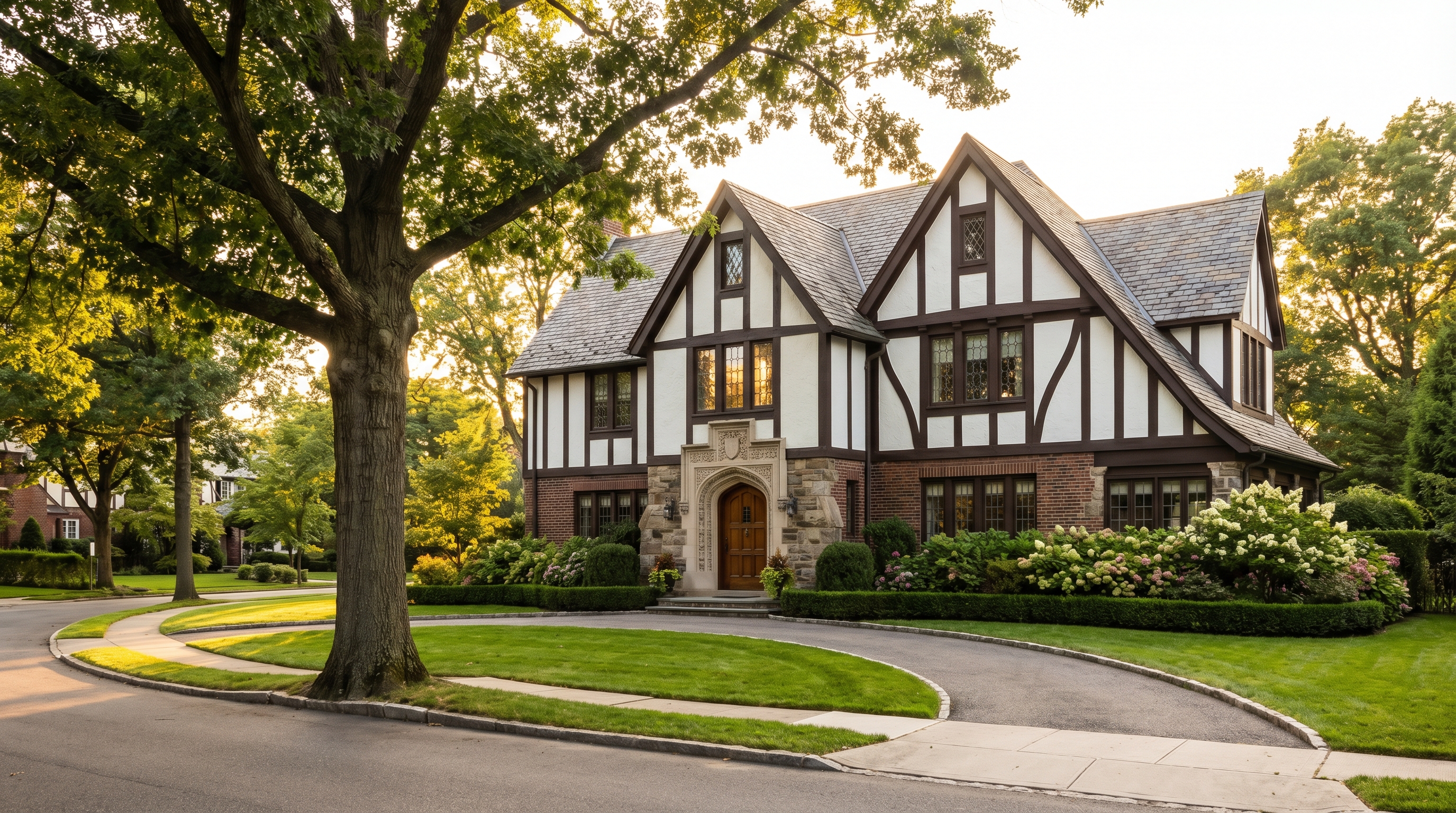 A Tudor Revival house in Jamaica Estates, Queens with half-timbered upper story, brick and stone facade, leaded casement windows, ornate entrance surround, and manicured foundation plantings on a curving residential street