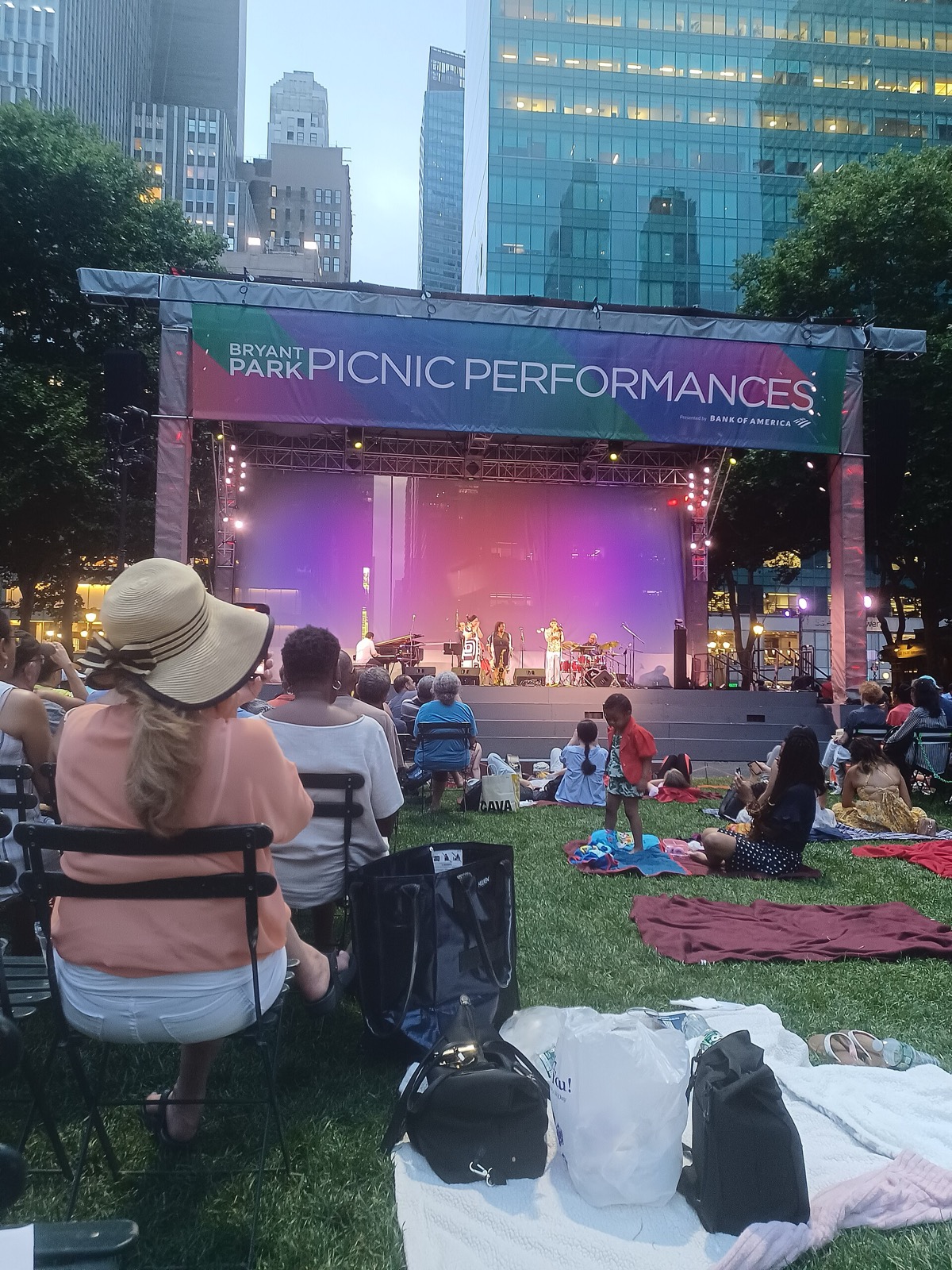 Bryant Park in Midtown Manhattan, showing the green lawn with visitors relaxing amid trees, the midtown skyline rising behind the park