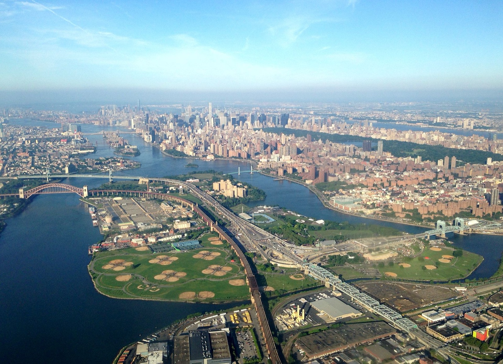 Aerial view of Randalls and Wards Islands from the east, showing the merged 480-acre island landmass, the athletic fields, RFK Triborough Bridge infrastructure, and the surrounding East River and Harlem River waterways
