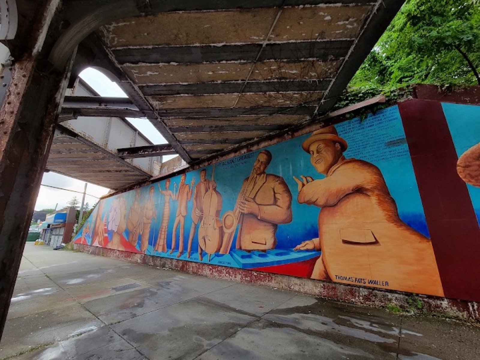 The St. Albans Greatest mural under the LIRR overpass at Linden Blvd and 180th Street, showing Thomas "Fats" Waller and fellow jazz legends painted in bold color against a vivid blue sky